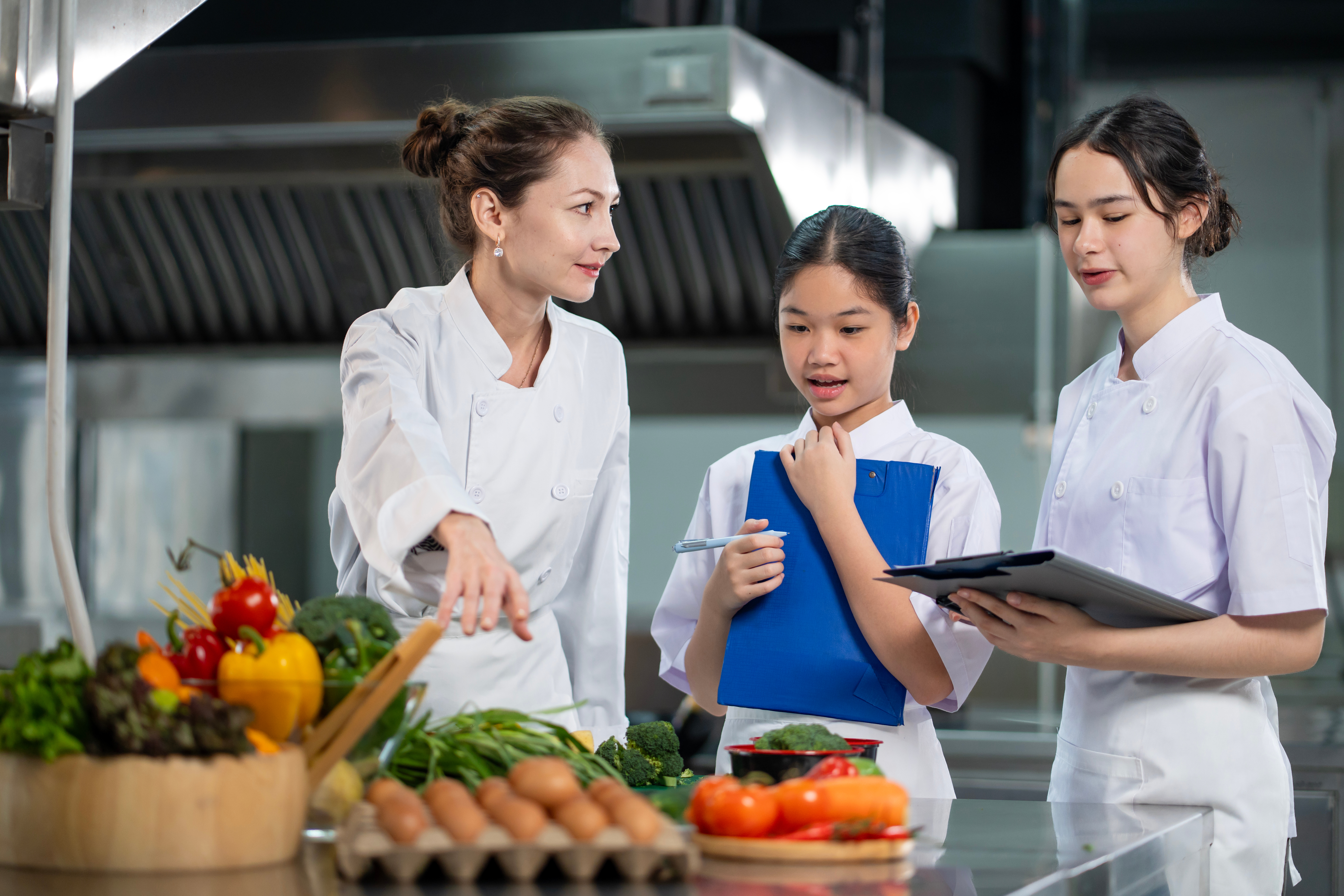 Three chefs in white coats engage in discussion in a professional kitchen. Fresh vegetables and eggs are on the counter.