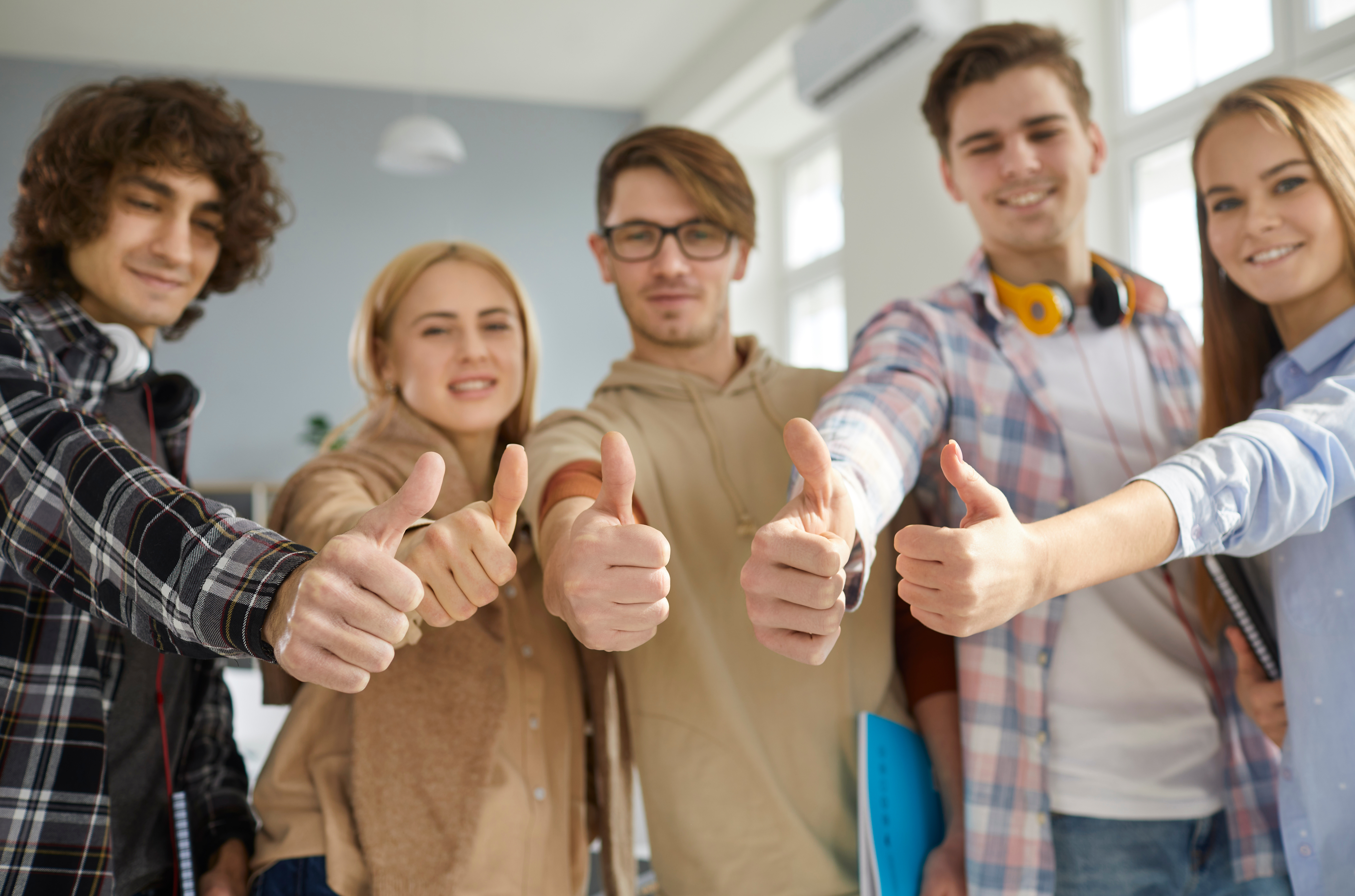 A group of five young adults stand in a bright room, all smiling and giving thumbs up.
