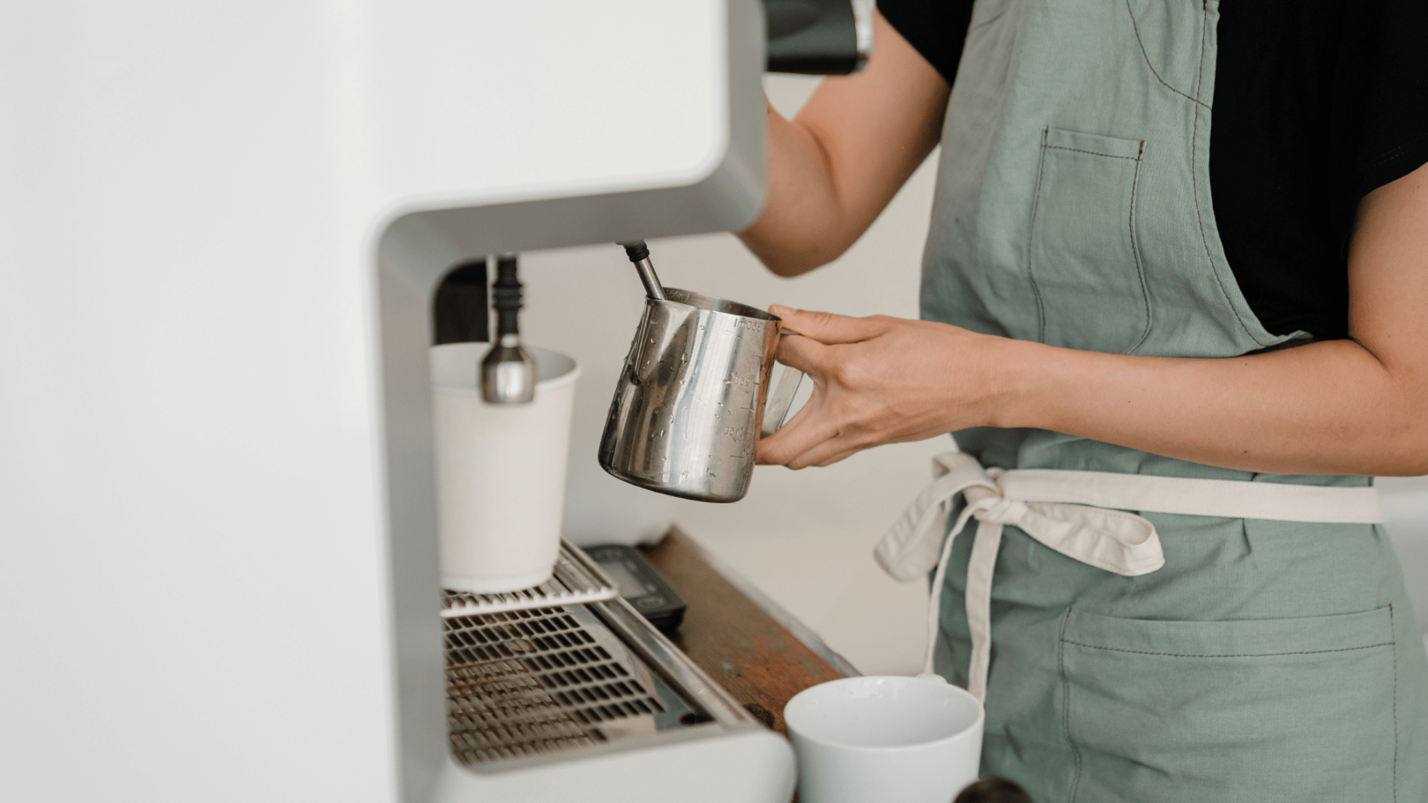 Barista in a green apron steams milk with a metal pitcher at an espresso machine