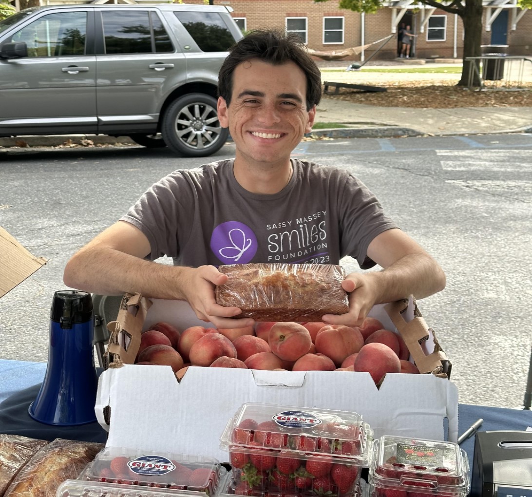 Smiling person holds a loaf of bread at an outdoor market stall with fresh peaches and strawberries